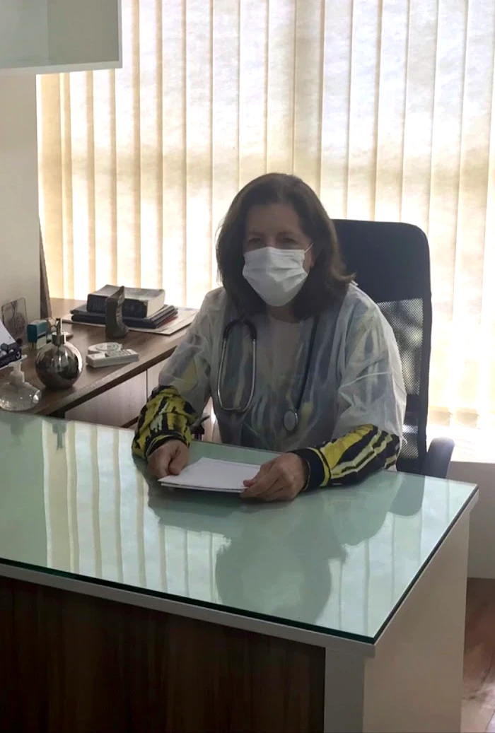 Dra. Marília à mesa de tampo de vidro do consultório, de jaleco, estetoscópio e máscara, com a persiana vertical clara atrás dela.