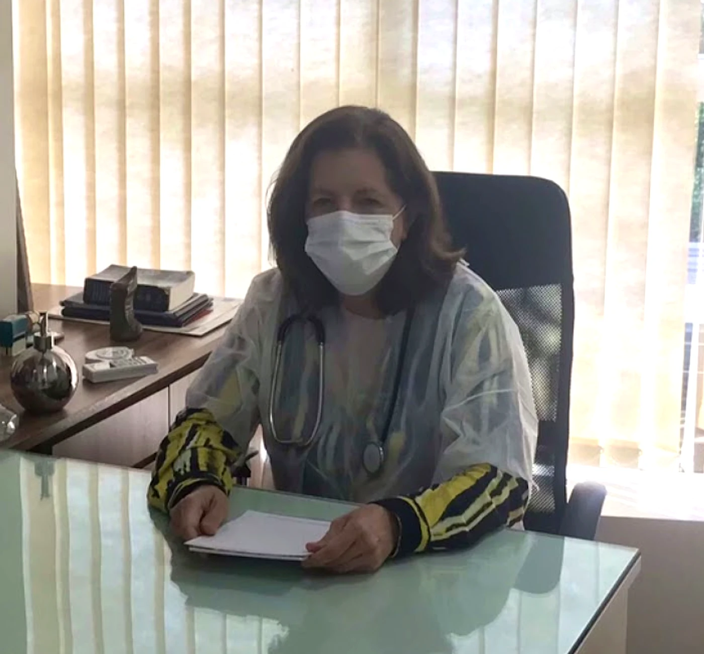 Dra. Marília sentada à mesa de vidro do consultório, de jaleco, máscara e estetoscópio no pescoço, com as mãos sobre um bloco de papel e a persiana clara atrás.